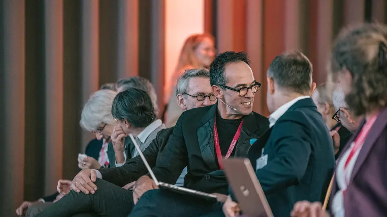 Side view of an audience; people are sitting in rows, chatting and laughing. A man turns around, laughing, to the row behind him.