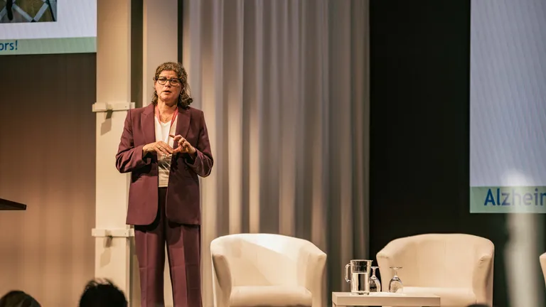 A woman in a wine-red trouser suit, her name is Prof. Charlotte Teunissen, stands on stage in front of an audience and speaks gesticulatingly.