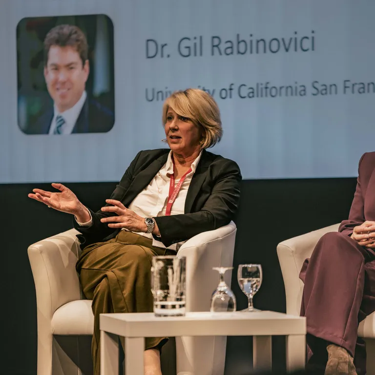Woman sitting on a white chair for a panel discussion, gesturing with her hands as she speaks.