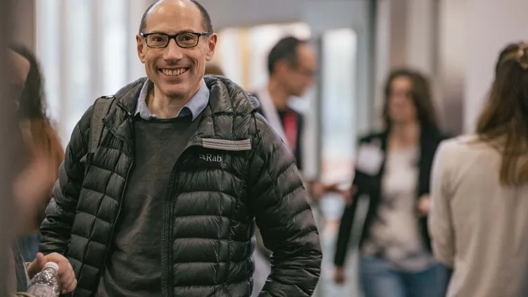 A man in a black jacket runs towards the camera, beaming; in the foreground and background, other people can be seen blurred.