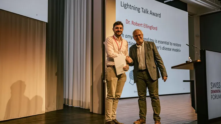 Two men are standing on a stage for an award ceremony; they shake hands and the award winner holds the certificate in his other hand.